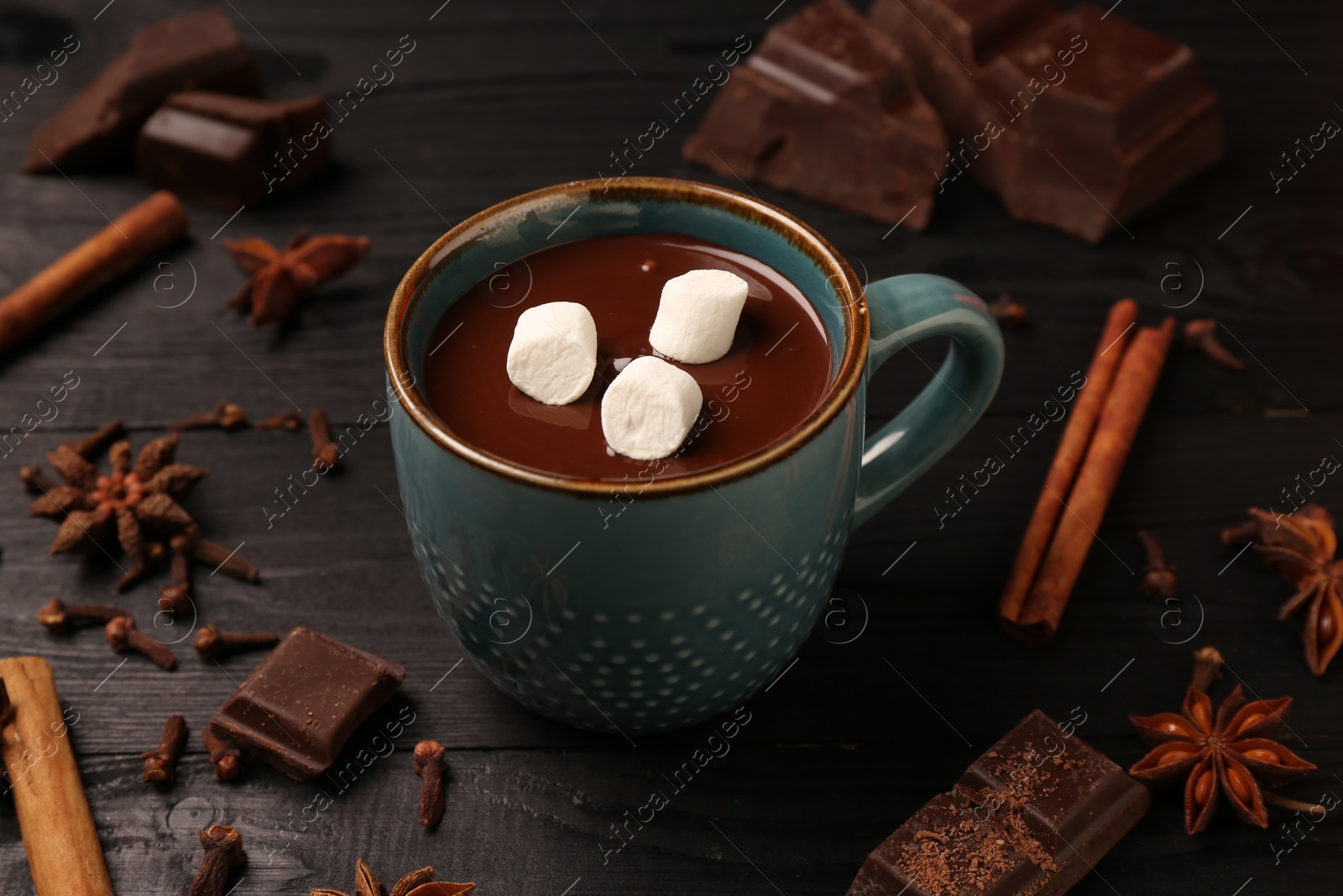 Photo of Tasty melted chocolate with marshmallows in cup and spices on dark wooden table, closeup