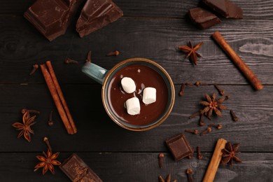 Photo of Tasty melted chocolate with marshmallows in cup and spices on dark wooden table, flat lay