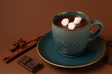 Photo of Tasty melted chocolate with marshmallows in cup and spices on brown background, closeup