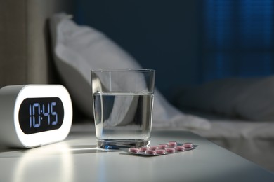 Photo of Insomnia treatment. Glass of water, pills and alarm clock on bedside table in bedroom at night