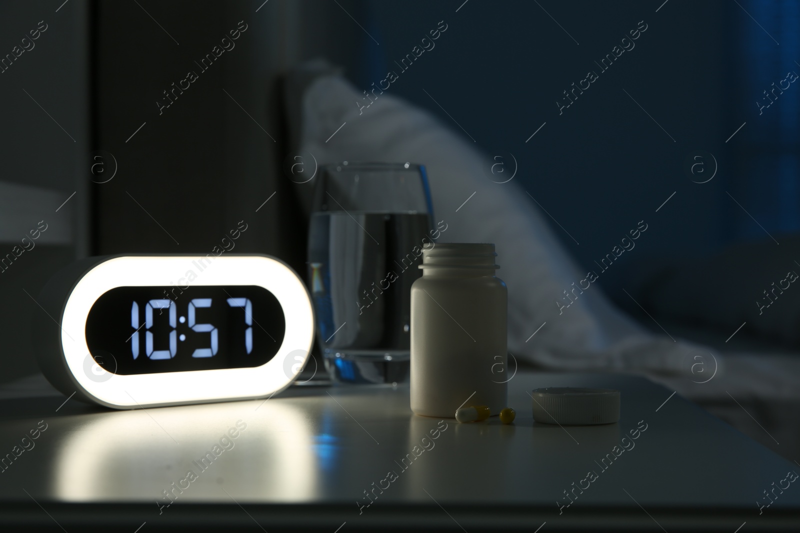 Photo of Insomnia treatment. Alarm clock, glass of water and pills on bedside table in bedroom at night