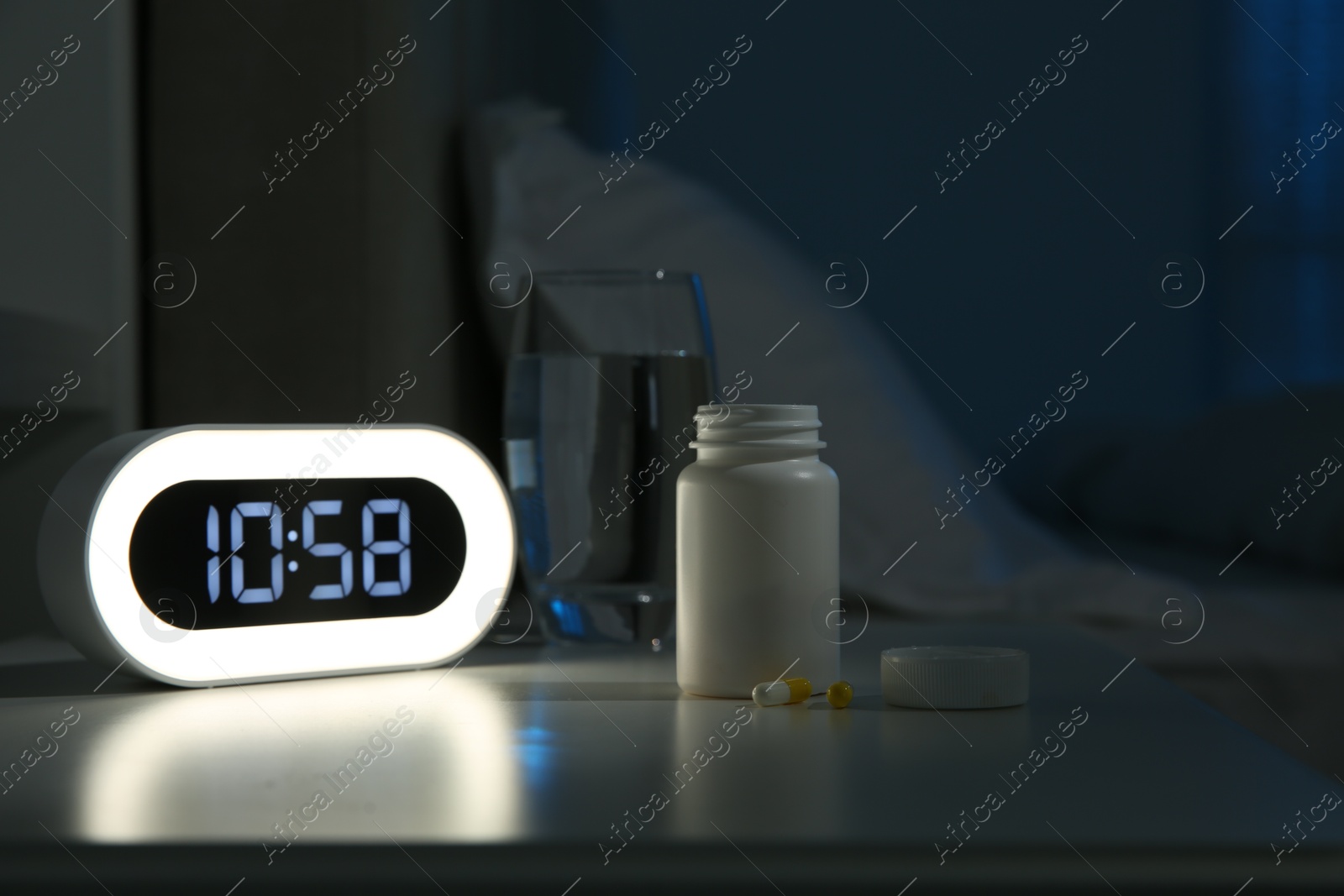 Photo of Insomnia treatment. Alarm clock, glass of water and pills on bedside table in bedroom at night