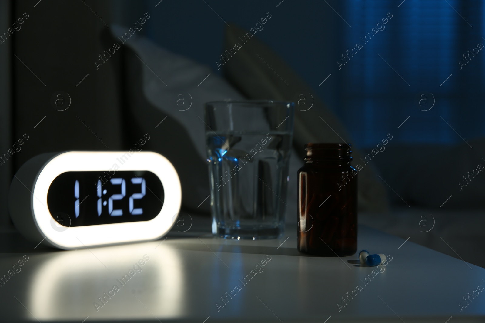 Photo of Insomnia treatment. Glass of water, pills and alarm clock on bedside table in bedroom at night