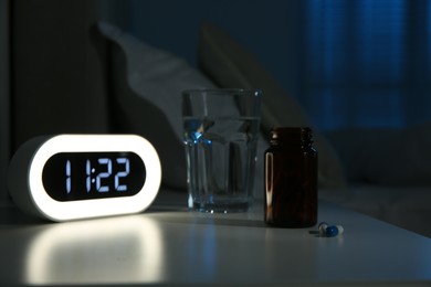 Insomnia treatment. Glass of water, pills and alarm clock on bedside table in bedroom at night