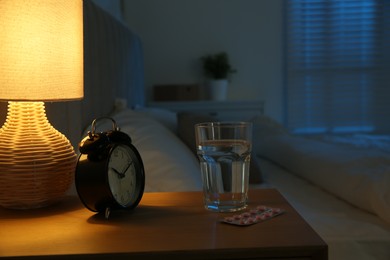 Photo of Insomnia treatment. Glass of water, pills and alarm clock on bedside table in bedroom at night