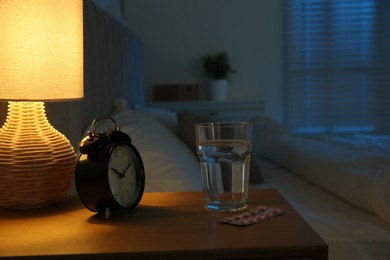 Insomnia treatment. Glass of water, pills and alarm clock on bedside table in bedroom at night