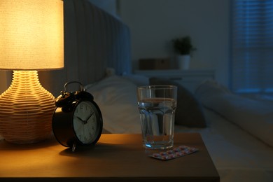 Insomnia treatment. Glass of water, pills and alarm clock on bedside table in bedroom at night