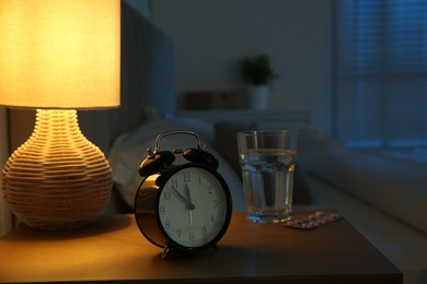 Photo of Insomnia treatment. Alarm clock, glass of water and pills on bedside table in bedroom at night