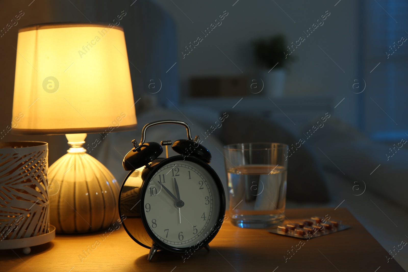 Photo of Insomnia treatment. Alarm clock, glass of water and pills on bedside table in bedroom at night