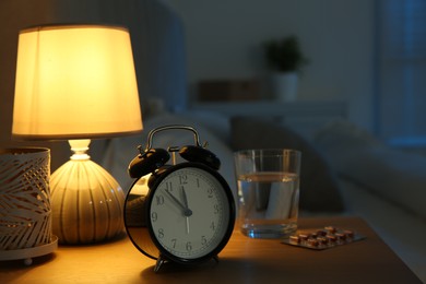 Photo of Insomnia treatment. Alarm clock, glass of water and pills on bedside table in bedroom at night