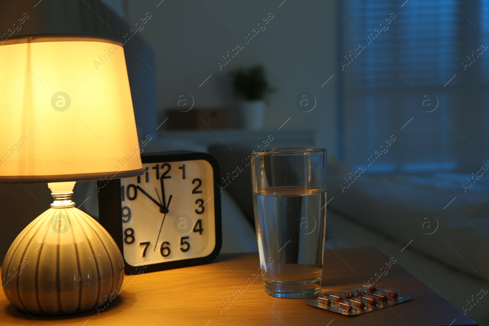 Photo of Insomnia treatment. Glass of water, pills and alarm clock on bedside table in bedroom at night