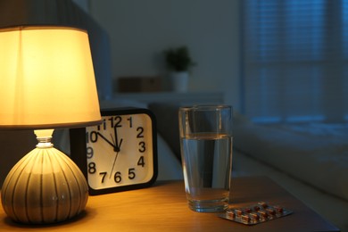 Insomnia treatment. Glass of water, pills and alarm clock on bedside table in bedroom at night