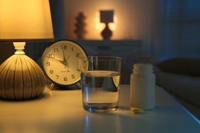 Insomnia treatment. Glass of water, pills and alarm clock on bedside table in bedroom at night