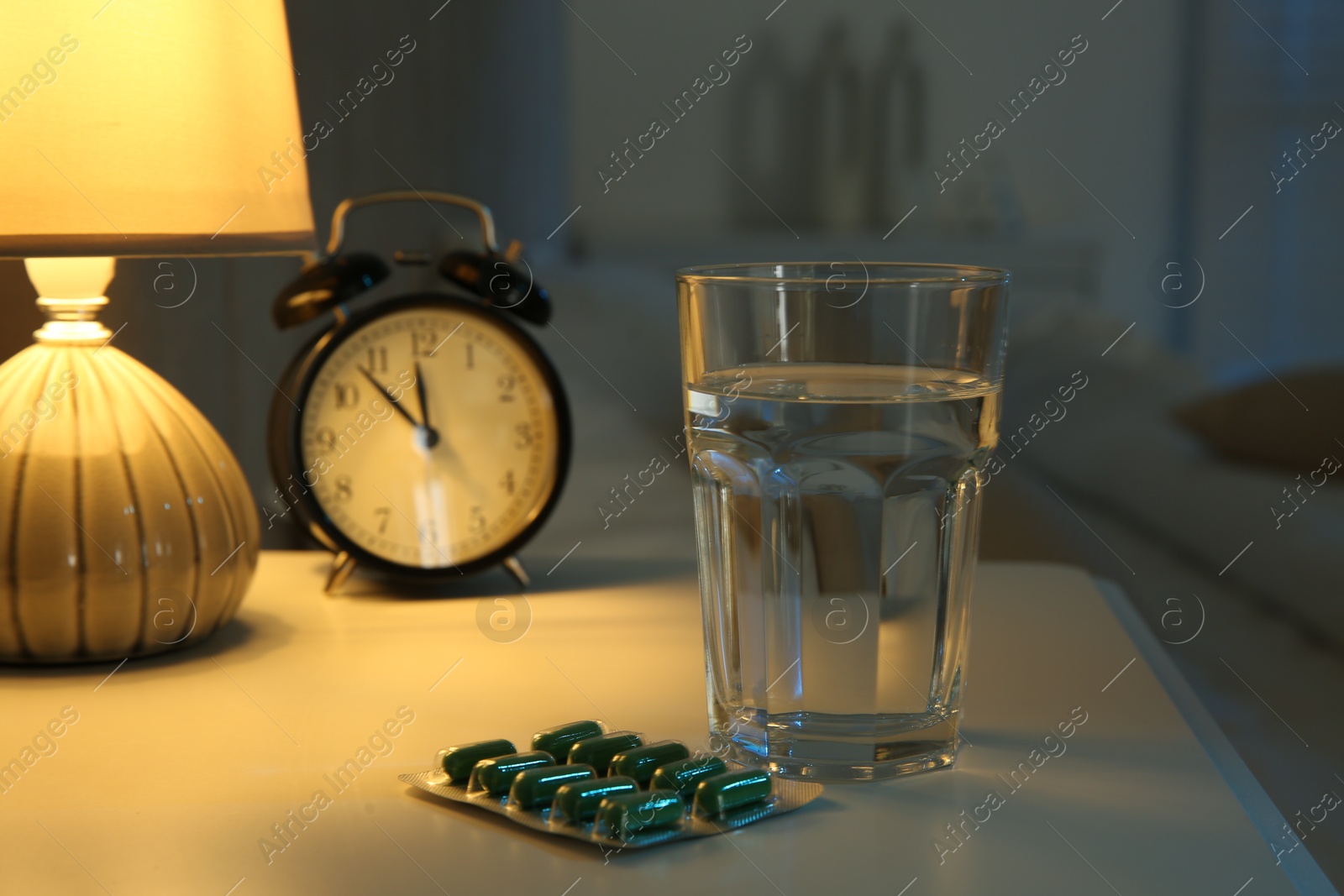 Photo of Insomnia treatment. Glass of water, pills and alarm clock on bedside table in bedroom at night