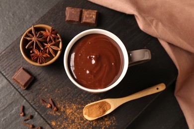 Tasty melted chocolate in cup and spices on dark textured table, top view