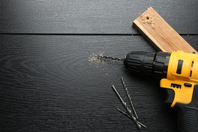 Cordless electric drill, bits and plank on black wooden table, flat lay. Space for text