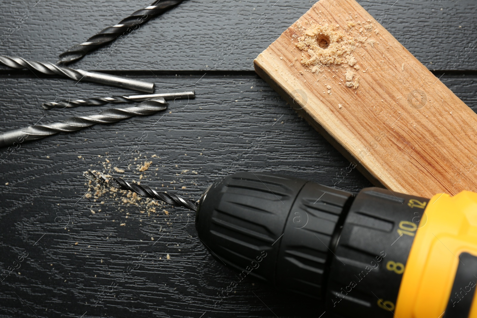 Photo of Cordless electric drill, bits and plank on black wooden table, flat lay