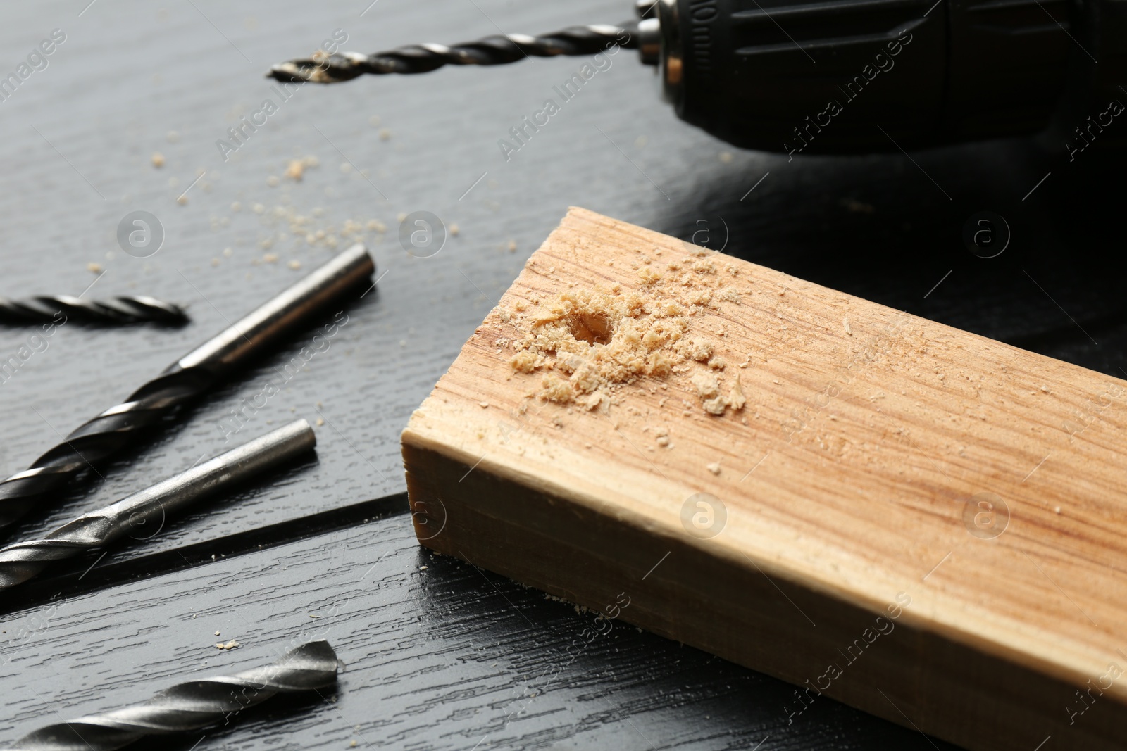 Photo of Cordless electric drill, bits and plank on black wooden table, closeup