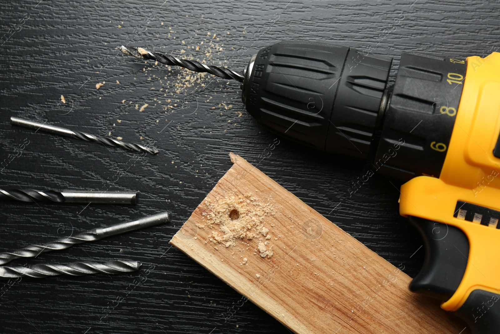 Photo of Cordless electric drill, bits and plank on black wooden table, above view