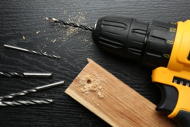 Cordless electric drill, bits and plank on black wooden table, above view