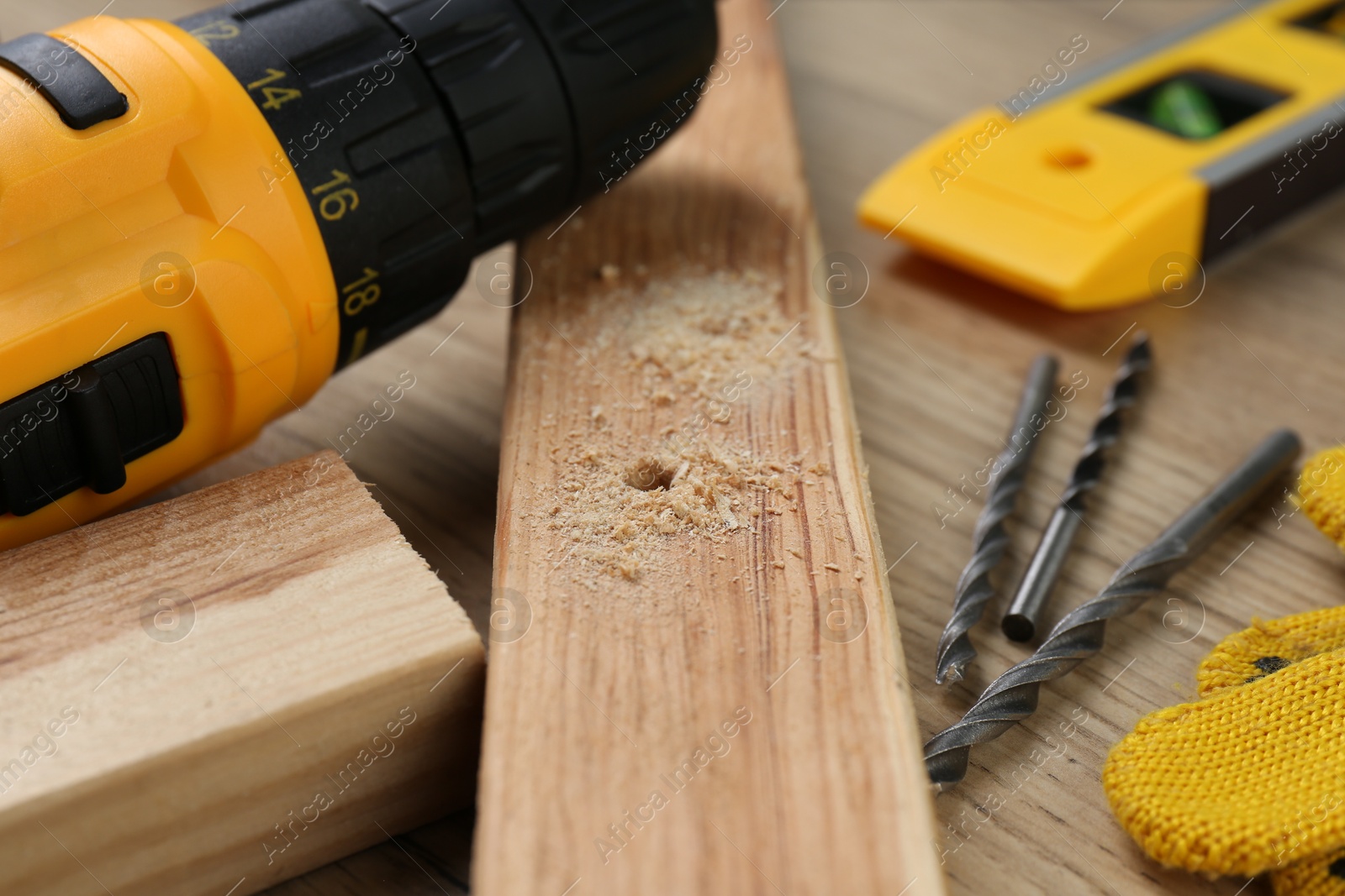 Photo of Cordless electric drill, bits, holed plank and sawdust on wooden table, closeup