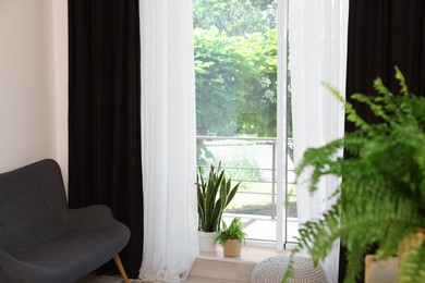 Photo of Shelving unit, sofa, green houseplants, pouffe and elegant curtains in room. Interior design