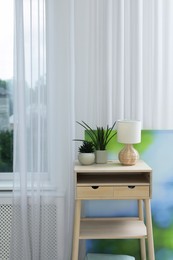 Photo of Nightstand, houseplants and lamp near window with beautiful curtain in room. Interior design