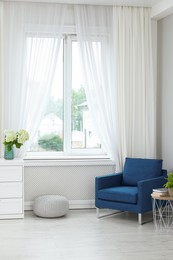 Soft armchair, side table, flowers and window with curtains in room. Interior design