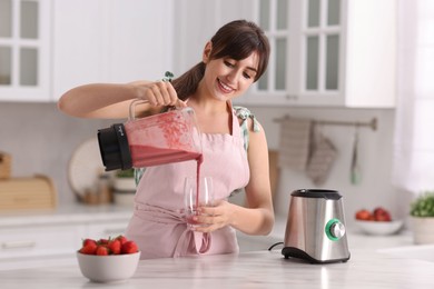 Photo of Young woman pouring fresh smoothie from blender cup into glass at white marble table in kitchen
