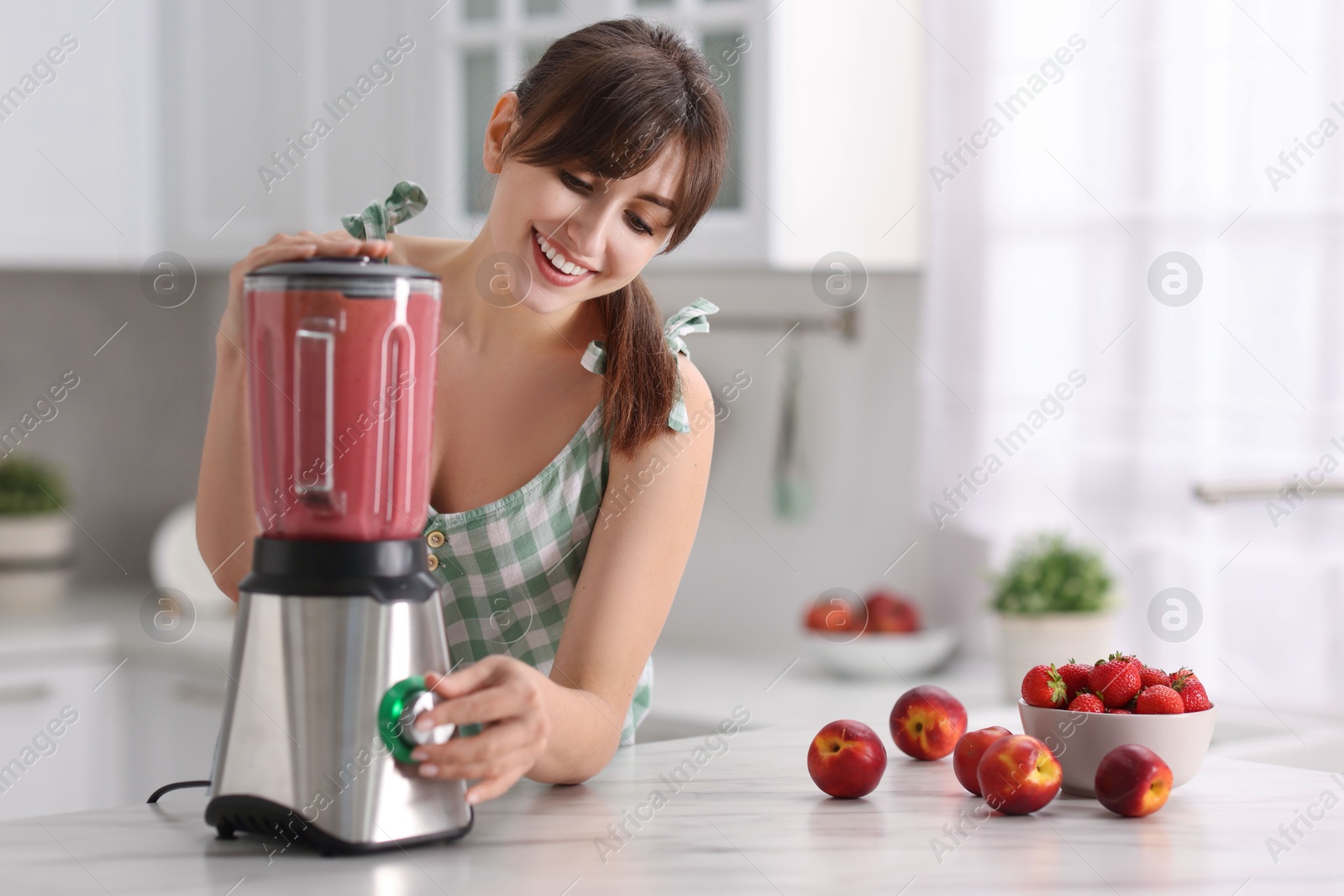 Photo of Young woman making delicious smoothie with blender at white marble table in kitchen