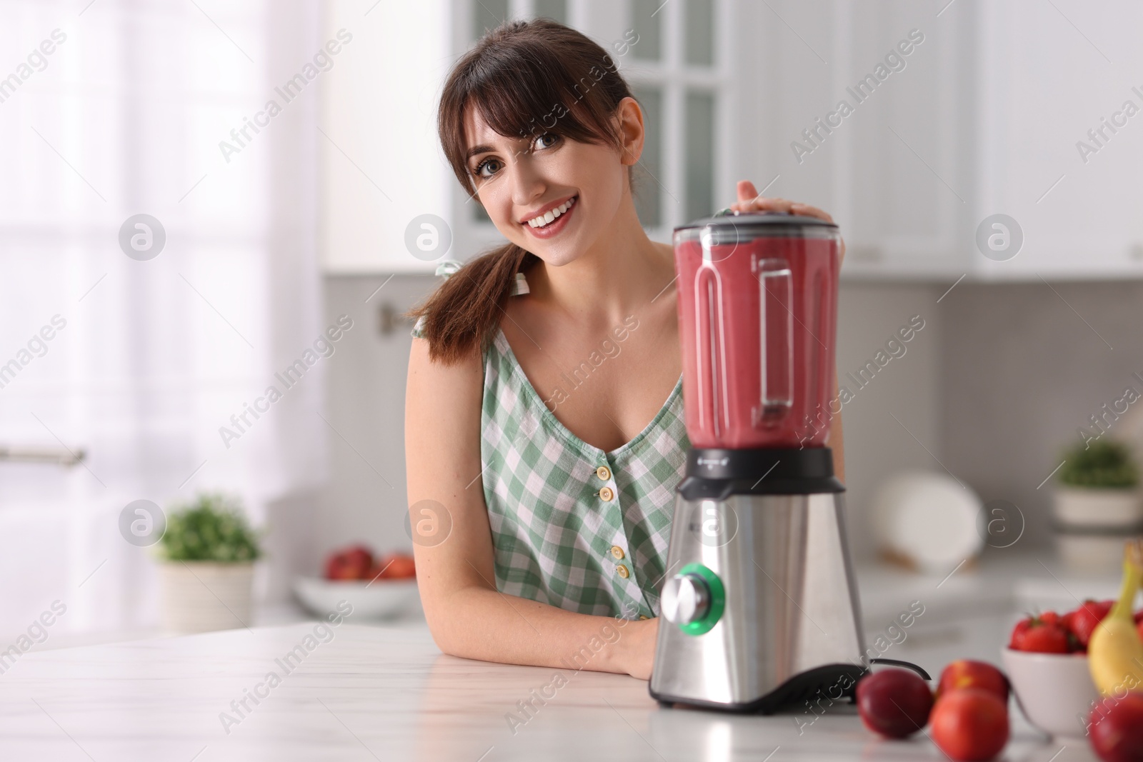 Photo of Young woman making delicious smoothie with blender at white marble table in kitchen