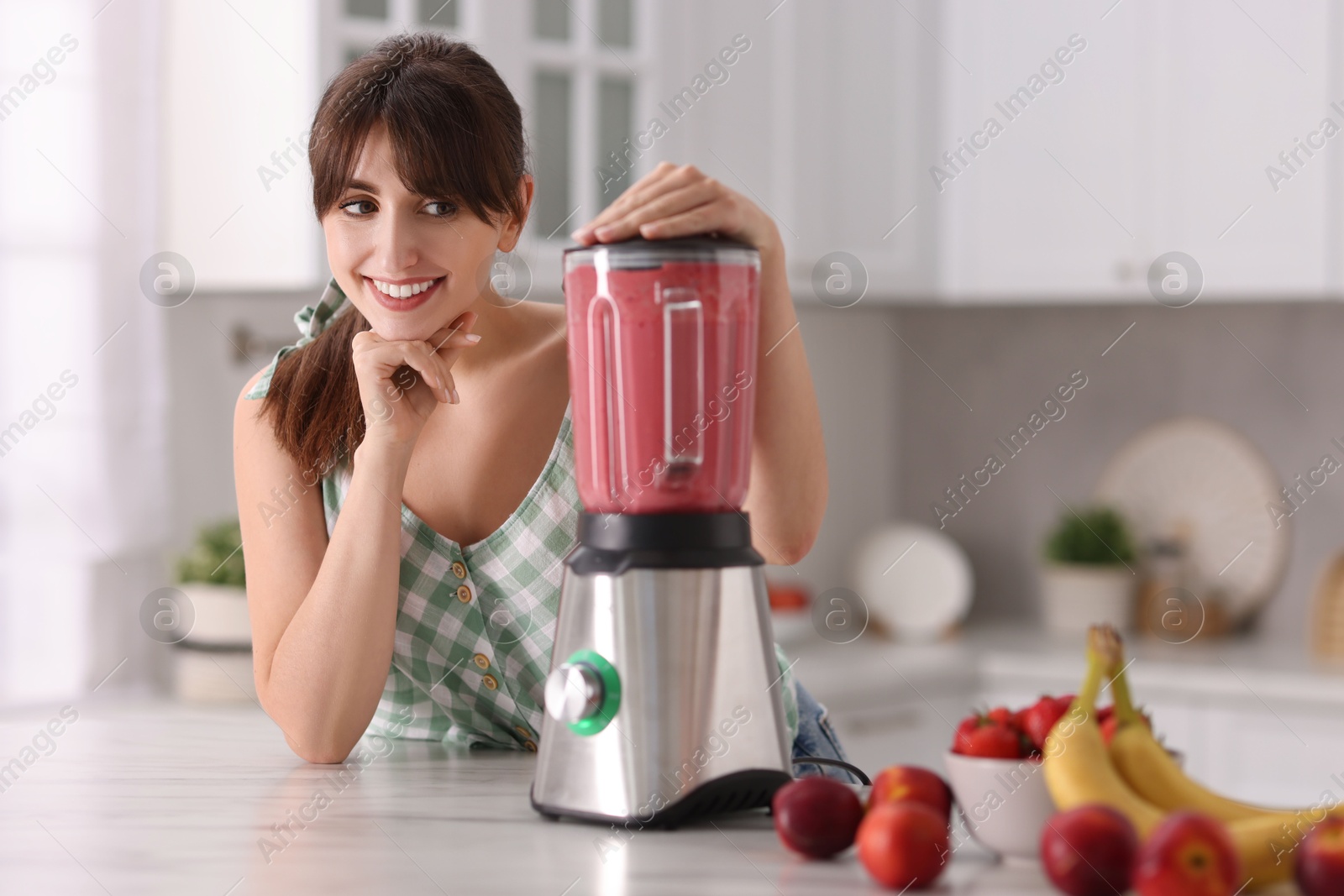 Photo of Young woman making delicious smoothie with blender at white marble table in kitchen