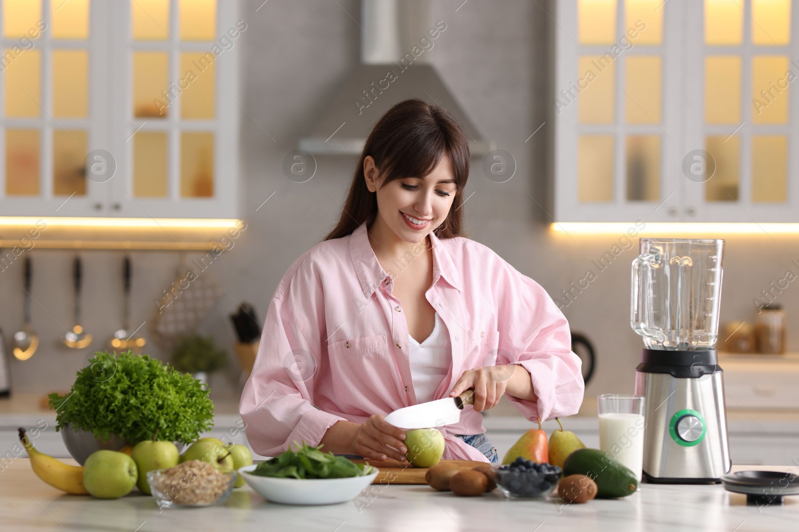 Photo of Young woman making delicious smoothie with blender at white marble table in kitchen