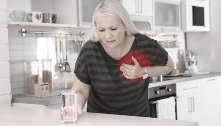 Image of Heart attack. Mature woman suffering from pain in chest and reaching for glass of water indoors. Banner design