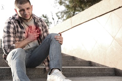 Image of Heart attack. Young man suffering from pain in chest on stairs outdoors