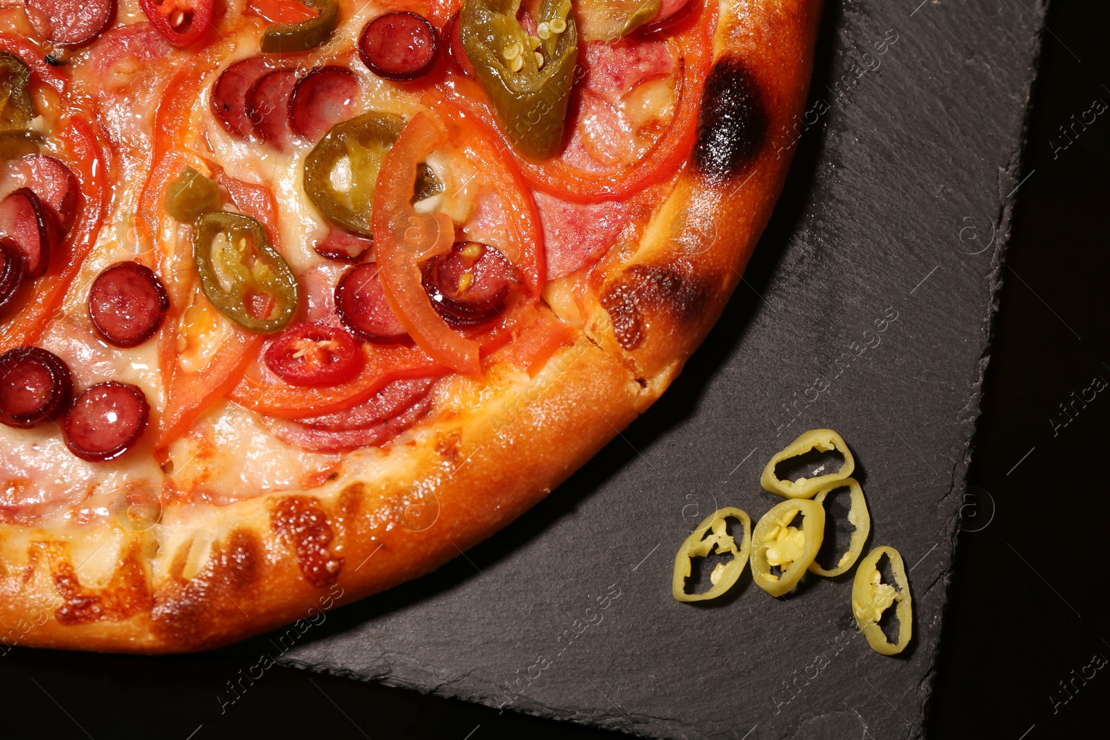 Photo of Delicious pizza Diablo and peppers on slate board against black background, top view