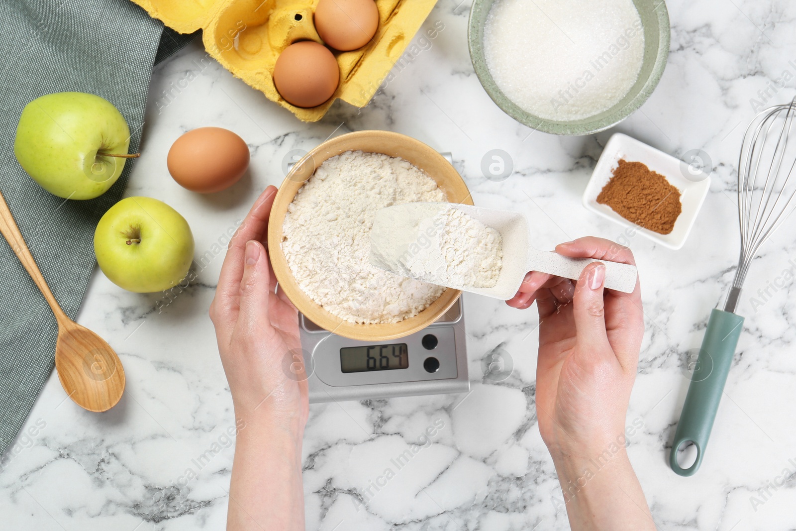 Photo of Woman adding flour into bowl on kitchen scale at white marble table, top view