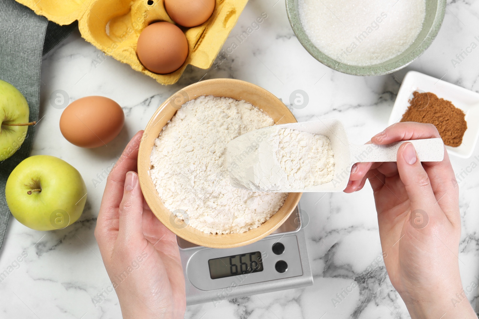 Photo of Woman adding flour into bowl on kitchen scale at white marble table, top view