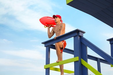 Handsome lifeguard with life buoy on watch tower against sky