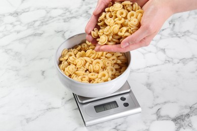 Woman adding pasta into bowl on kitchen scale on white marble table, closeup