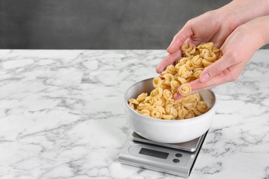 Photo of Woman adding pasta into bowl on kitchen scale on white marble table, closeup. Space for text