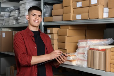 Smiling young man with clipboard near cardboard boxes in auto store