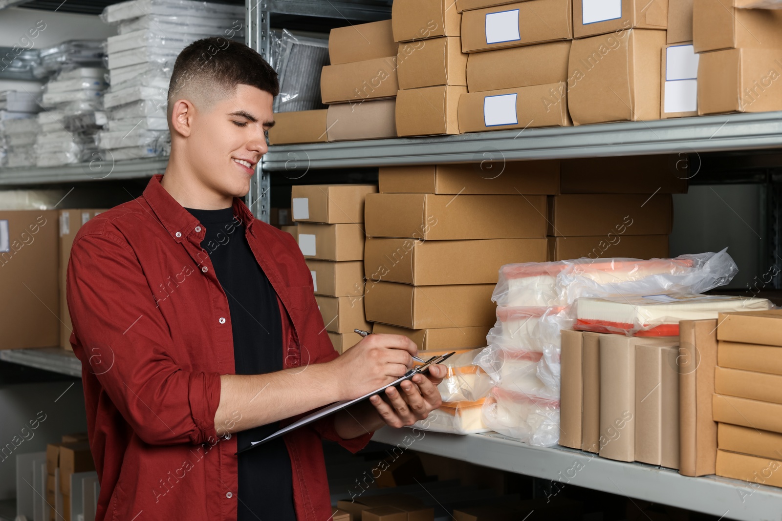 Photo of Young man with clipboard taking notes near cardboard boxes in auto store