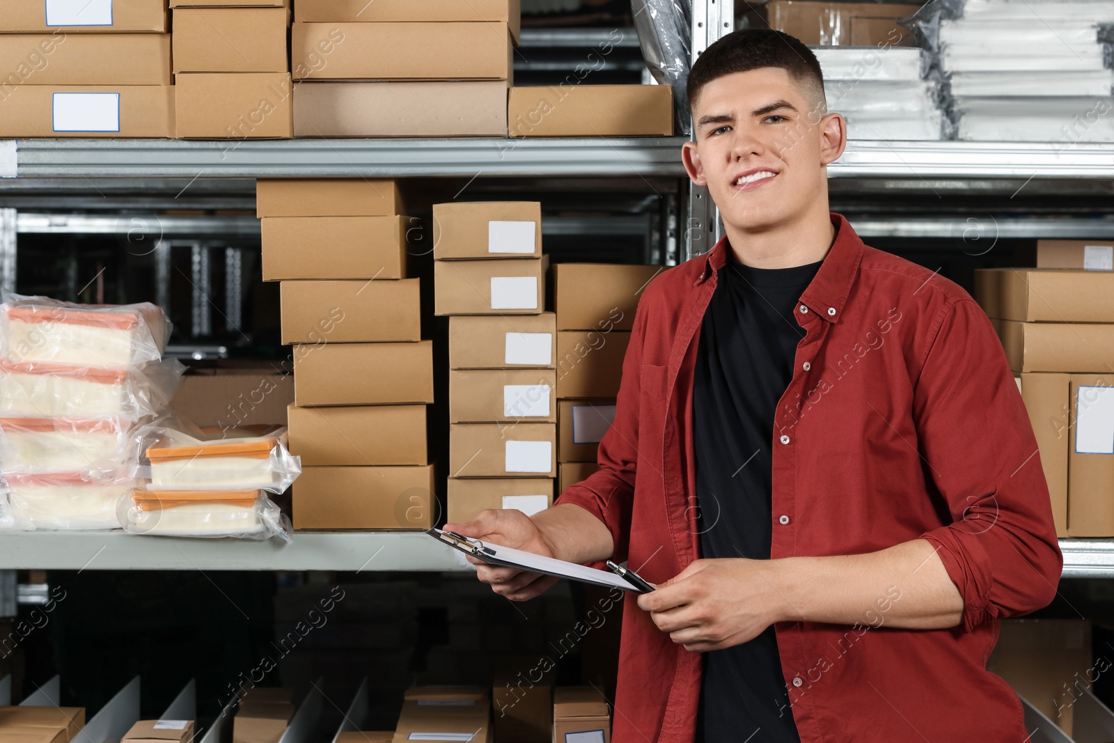 Photo of Smiling young man with clipboard near cardboard boxes in auto store