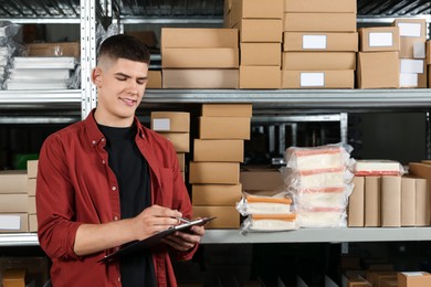 Smiling young man with clipboard near cardboard boxes in auto store