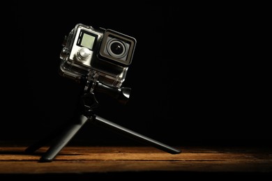 Photo of Modern action camera and tripod on wooden surface against black background. Space for text