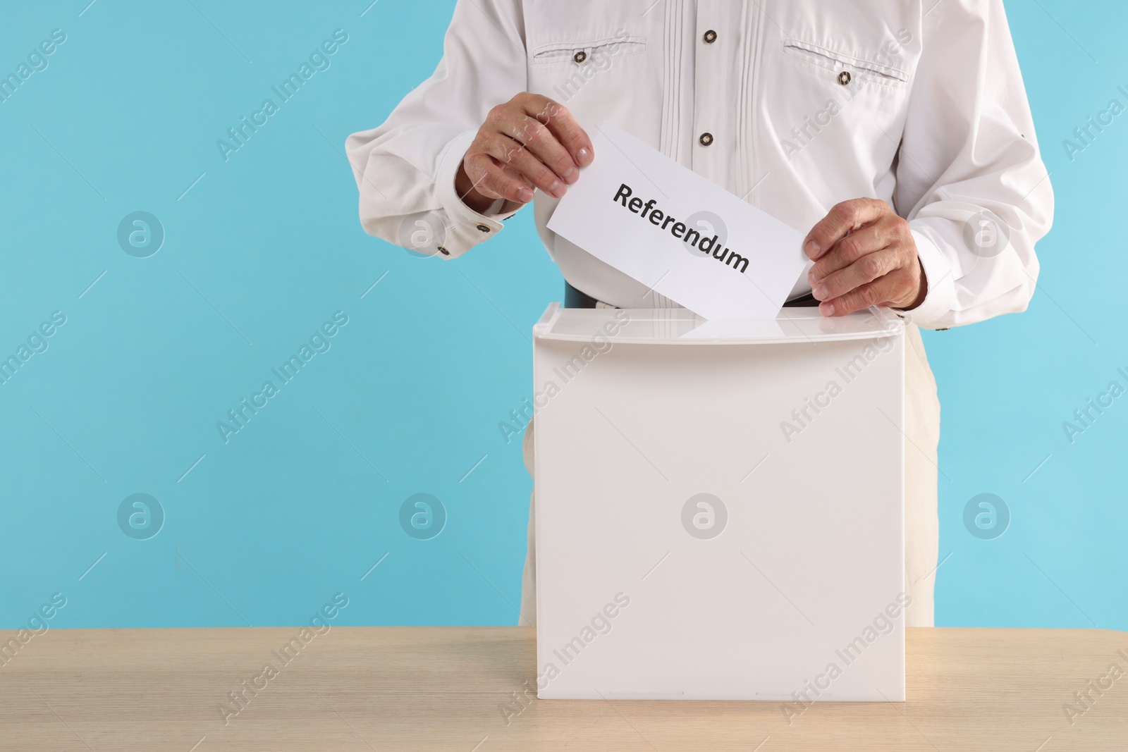 Photo of Referendum. Man putting his vote into ballot box at wooden table against light blue background, closeup