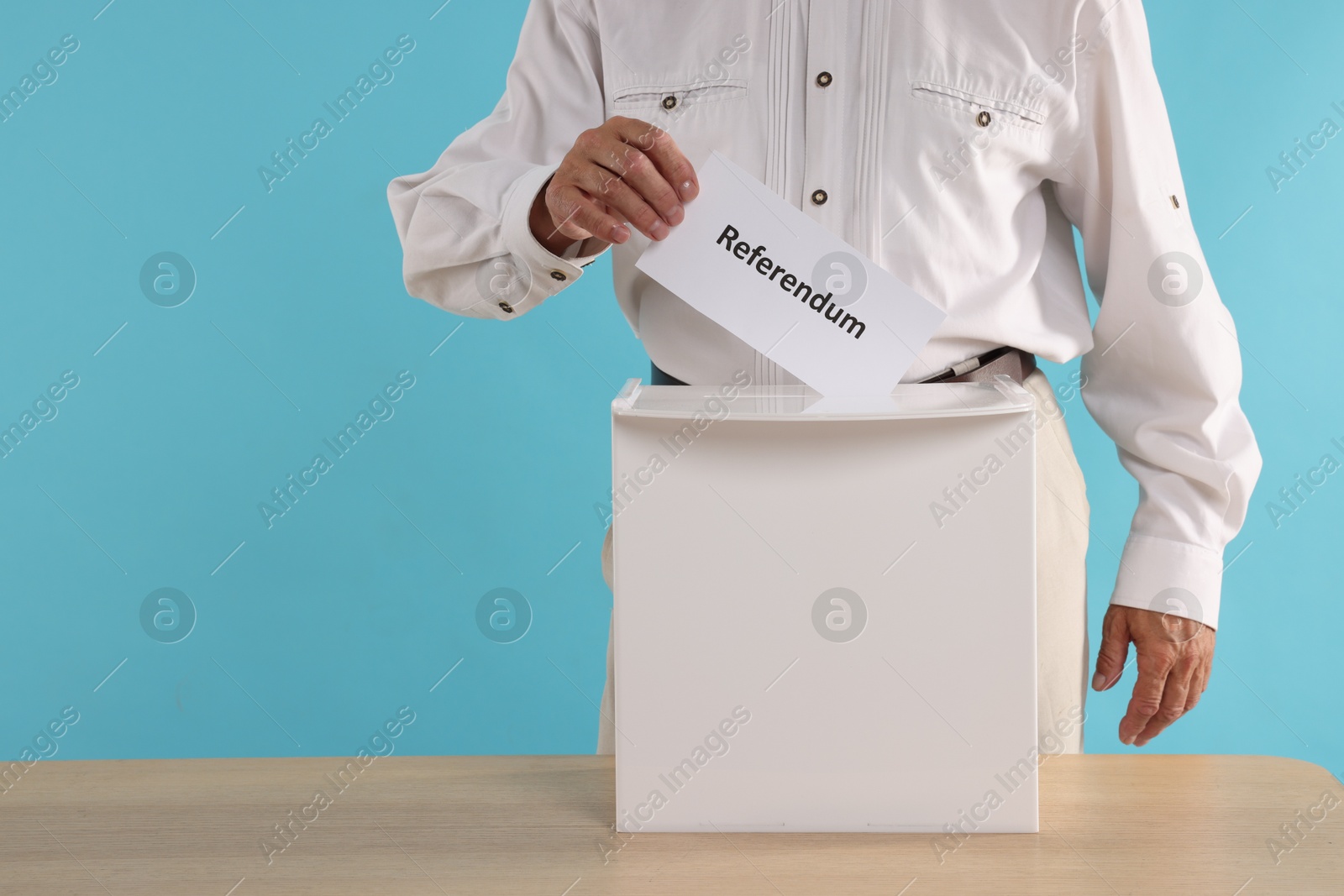 Photo of Referendum. Man putting his vote into ballot box at wooden table against light blue background, closeup