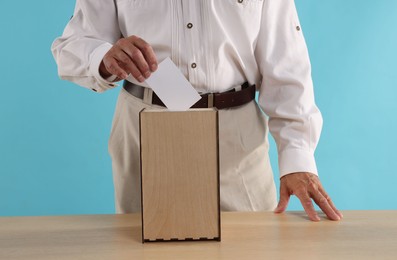 Photo of Referendum. Man putting his vote into ballot box at wooden table against light blue background, closeup