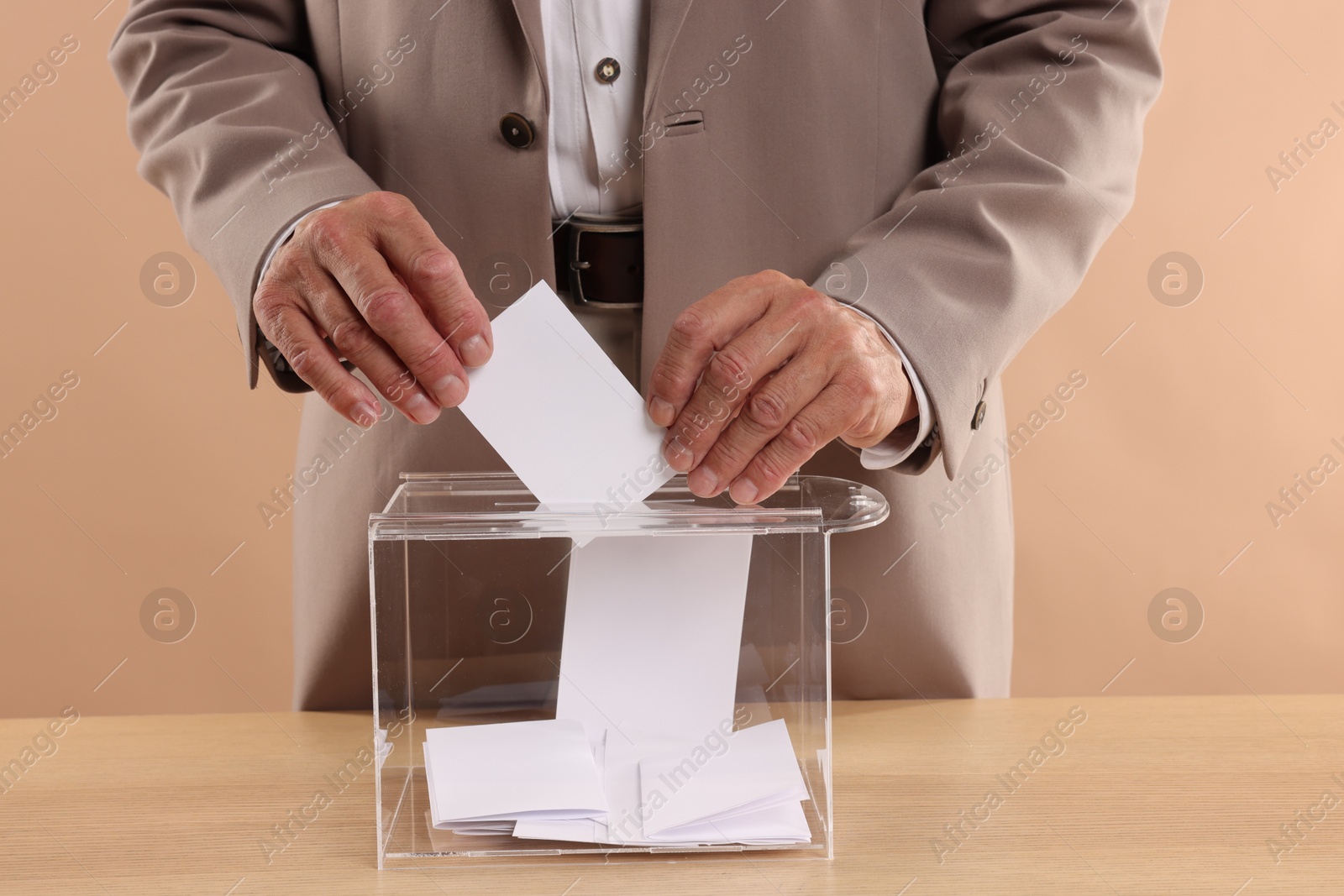 Photo of Referendum. Man putting his vote into ballot box at wooden table against beige background, closeup